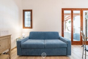 Une pièce avec un canapé bleu, un meuble en bois, une petite table avec une lampe et une fenêtre avec des barreaux.