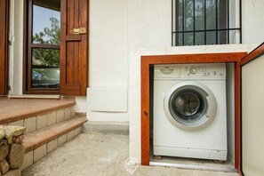 Une petite machine à laver blanche, placée dans un coffrage en bois, se trouve sur un sol en béton, à côté d’un escalier en pierre menant à une porte également encadrée de bois.