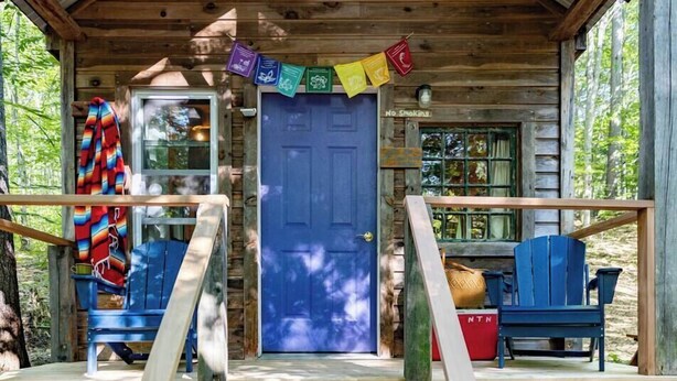 Une cabane avec une porte bleue, une véranda avec des marches en bois et deux chaises Adirondack bleues.