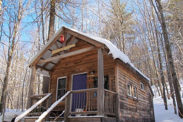 A wooden cabin with a porch and a blue door, surrounded by a snowy forest. - "Robins Nest" Off Grid Solar Powered Eco Cabin (Denmark)