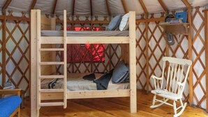 Een houten stapelbed met een ladder, een witte schommelstoel en een plank met boeken en een hoed.