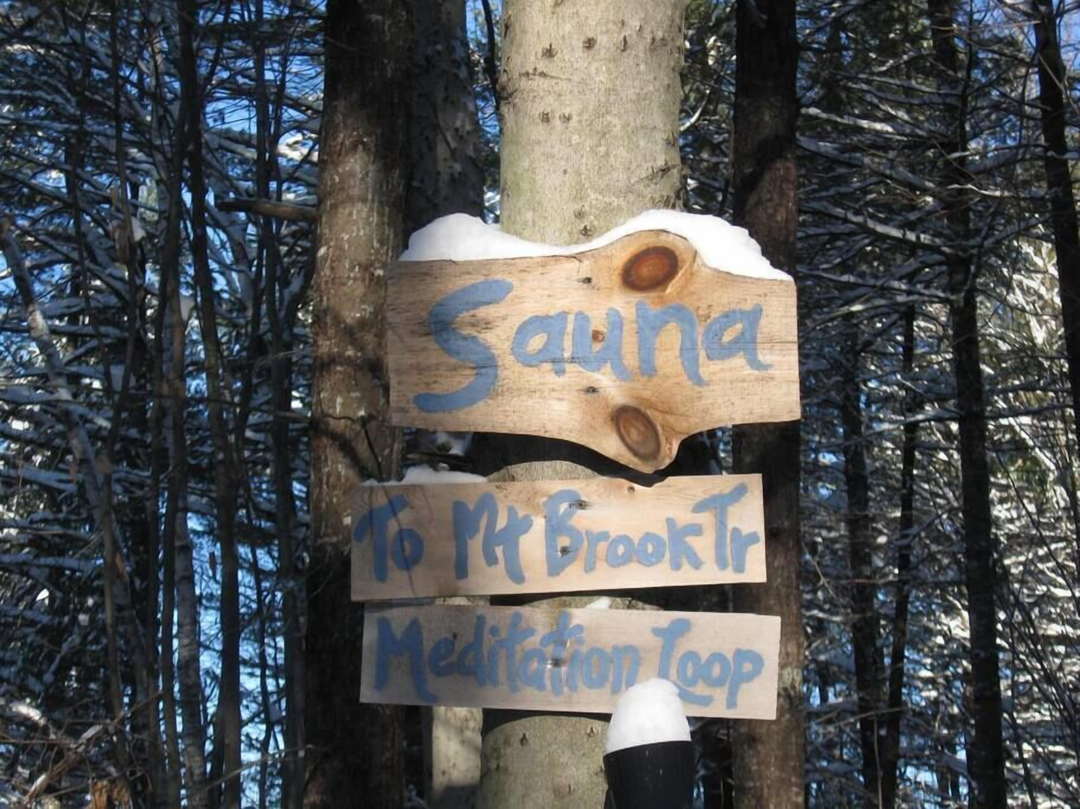 A wooden sign with 'Sauna' and 'Mt. Brook Tr Meditation Loop' written on it, attached to a tree in a snowy forest.