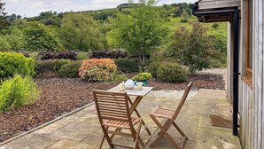 An outdoor patio with a table and two wooden chairs, surrounded by a garden with various plants and a view of rolling hills in the background.