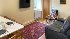 A modern living room with a flat-screen TV, a wooden door, a red patterned rug, a black leather sofa, and a dining area with a table and chairs.