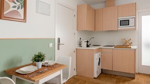 A compact kitchen with wooden cabinets, a microwave, a washing machine, and a dining area with a table and chairs. - Sol Studio II – In the City Center (Madrid)