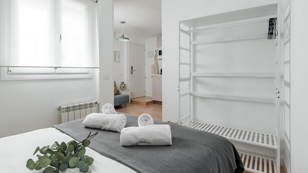 A modern bedroom with a bed, a radiator, a window, and a white shelving unit. - Sol Studio IV – In the City Center (Madrid)