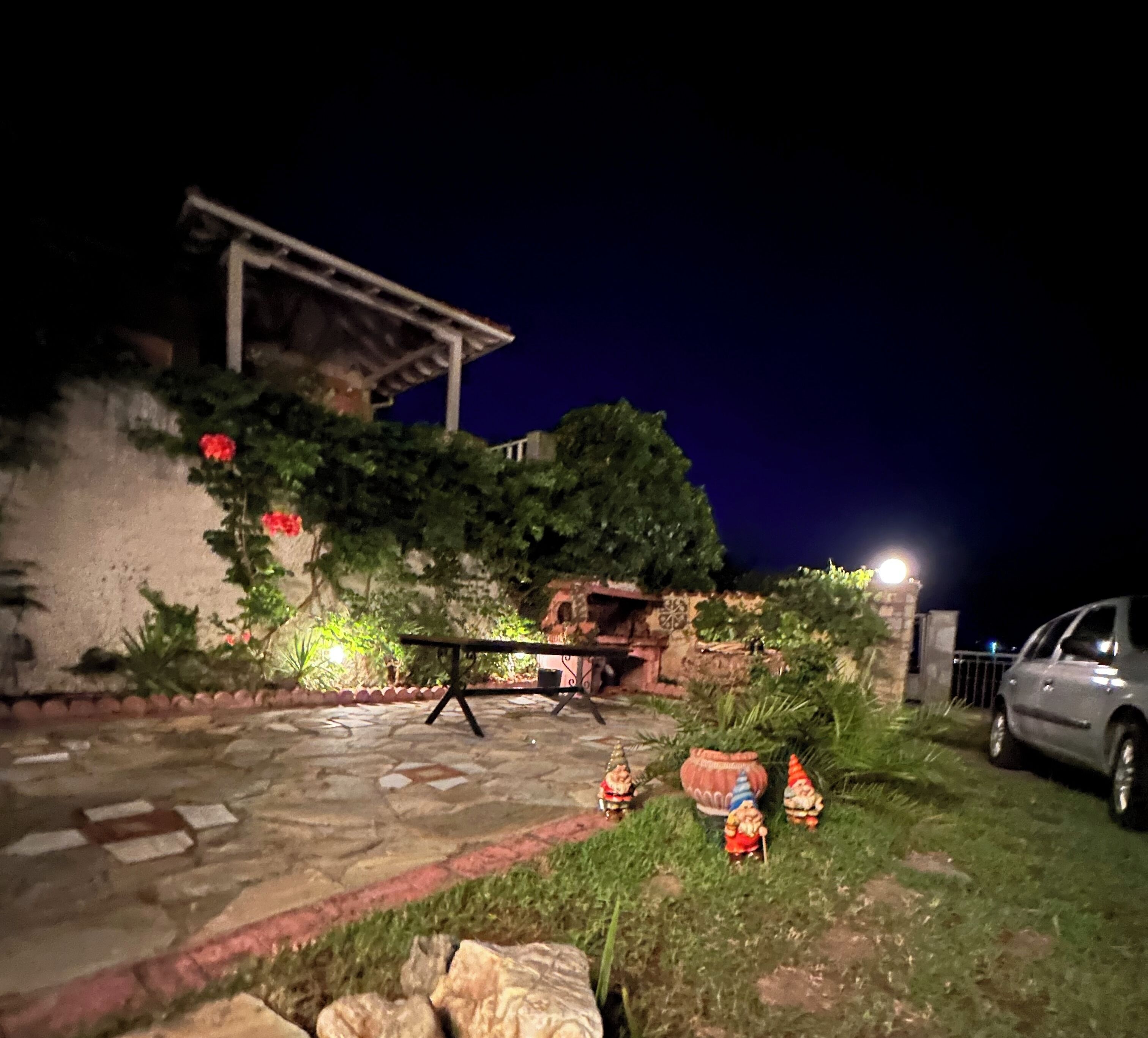 A stone patio with a table and chairs, surrounded by greenery and a car parked nearby.