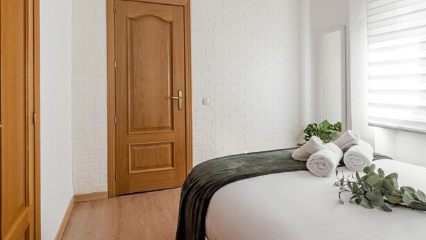 Un dormitorio con puerta de madera, paredes de ladrillo blanco y un patrón decorativo, cama con manta verde, toallas enrolladas y una planta.