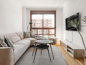 Ein modernes Wohnzimmer mit einer Couch, einem Couchtisch, einem Fernseher und einem großen Fenster mit Blick auf Gebäude.