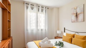 A neatly arranged hotel room with a bed, a nightstand, a lamp, and a window with sheer curtains.