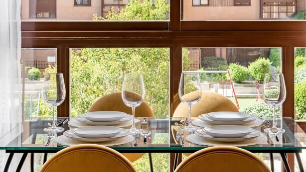 A dining table set with plates, glasses, and cutlery, positioned in front of a window with a view of a brick building and greenery.