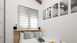 Een moderne slaapkamer met een bed, witte beddengoed, een mosterdkleurige plaids en een raam met zonwering.