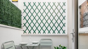 Een klein terras met een metalen tafel en twee stoelen, een plant in een pot en een groene, houten muur.