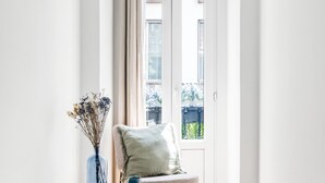 Een kamer met een raam, een stoel met een kussen en een vaas met gedroogde bloemen.
