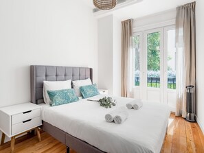 A modern bedroom with a grey upholstered headboard, white bedding, and a view of greenery through a large window. - Sol Apartment II - In the center of Madrid (Madrid)