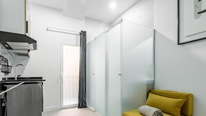 Una habitación moderna y minimalista con una lavadora gris, un sofá amarillo con una toalla y un ventanal corredero hacia un balcón.