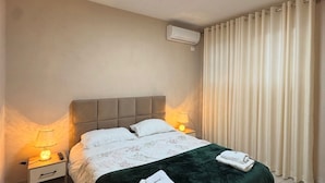 Un dormitorio ordenado con una cama, dos mesitas de noche, un aire acondicionado de pared y una ventana con cortinas.