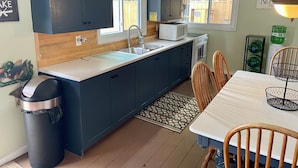 A compact kitchen with dark blue cabinets, a white countertop, a microwave, a sink, and a dining area with a table and chairs. - Ostrander's 3 Bedroom Cottage on Rice Lake (Harwood)