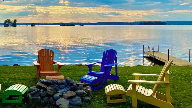 A lakeside view with a dock, a stone fire pit, and three Adirondack chairs in different colors. - Ostrander's 3 Bedroom Cottage on Rice Lake (Harwood)
