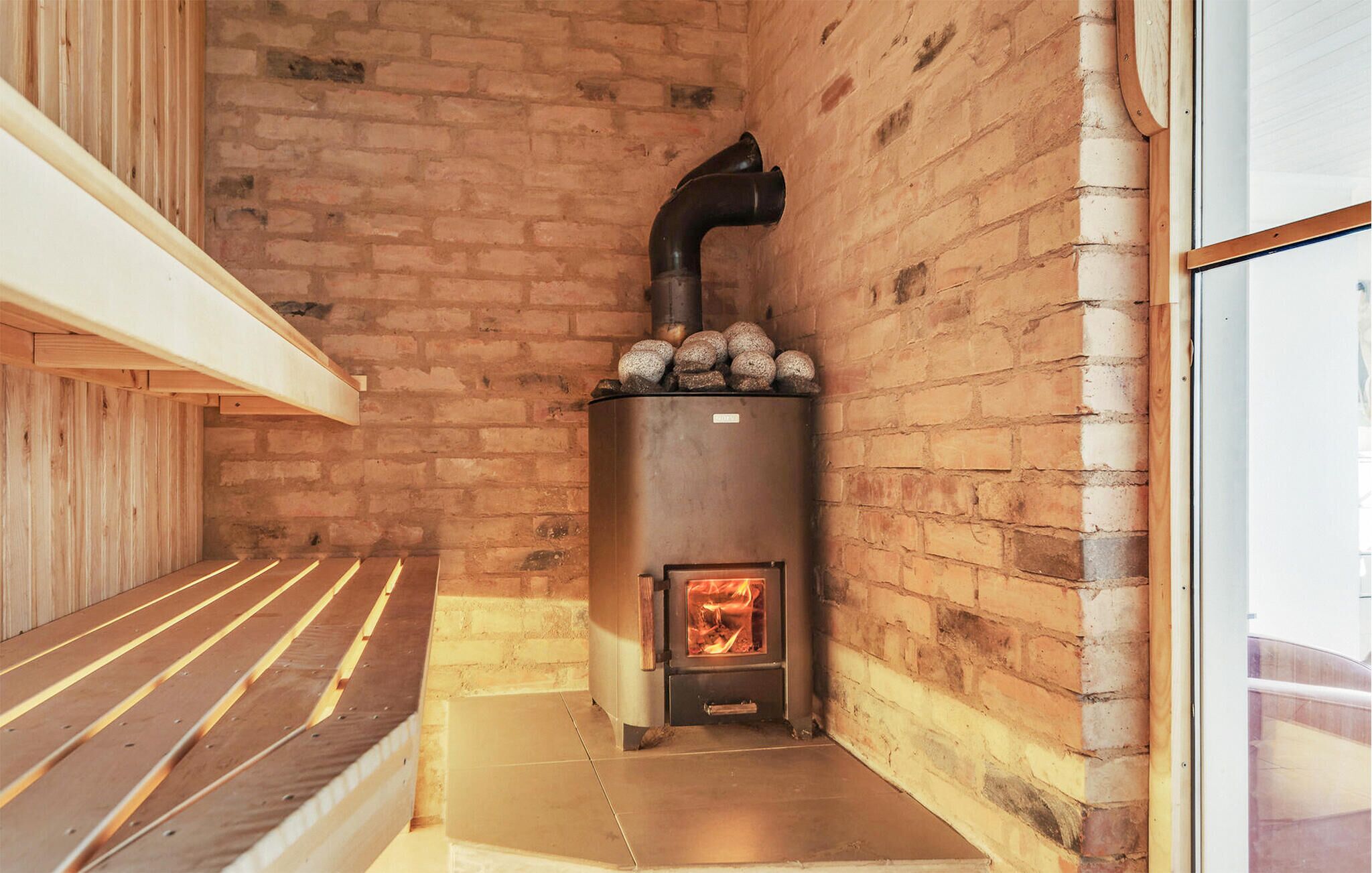 A wood-burning stove with a fire inside, set against a brick wall in a room with a wooden bench and a glass door.