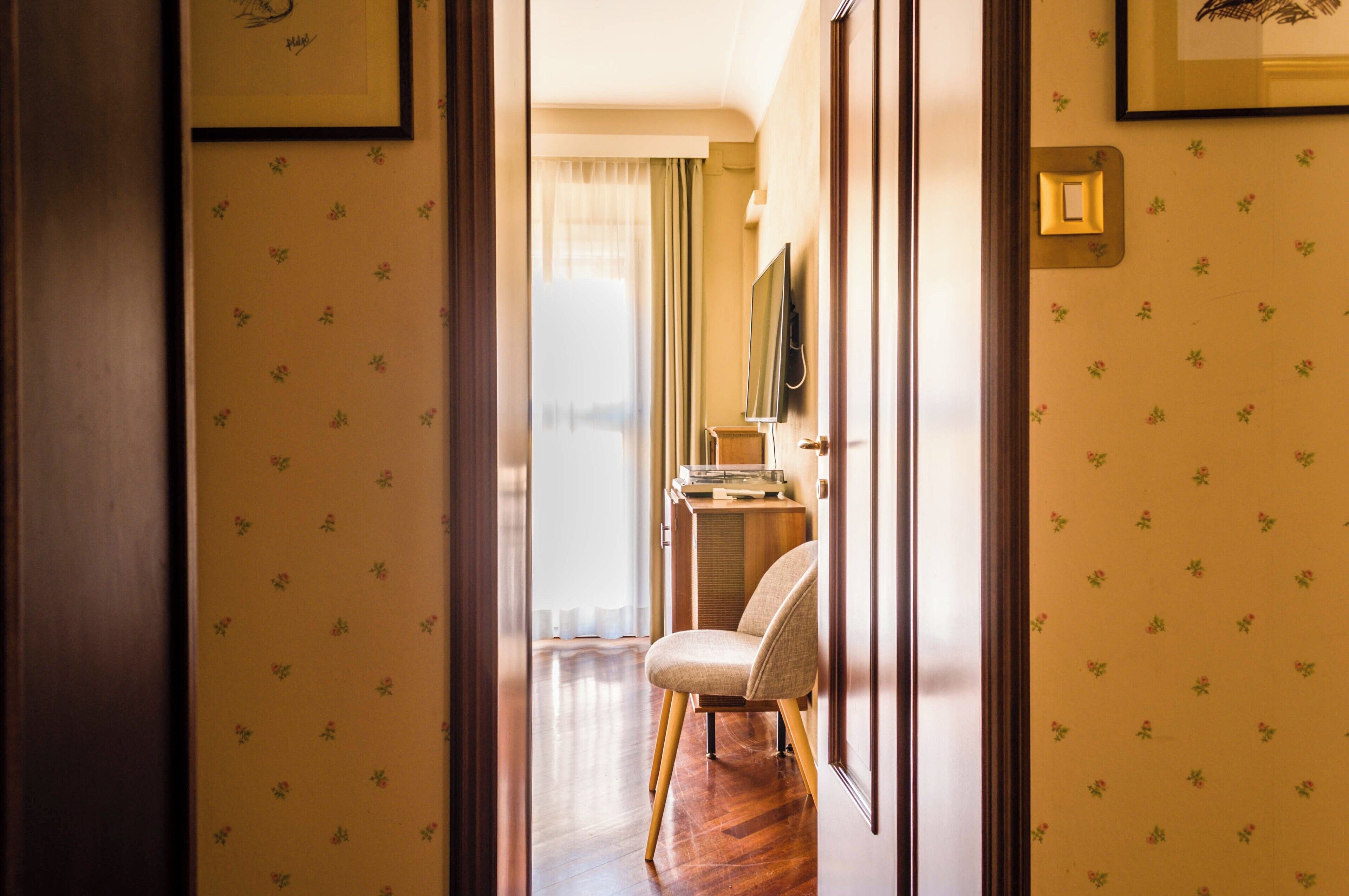 A hallway with floral wallpaper, a wooden floor, a chair, and a television on a stand.