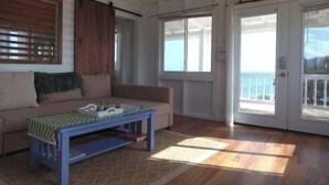 A room with a wooden floor, a beige sofa, a blue coffee table, and a window with blinds. - Barbuda Cottages 2 bed ( Green ) Beachfront Villa
 (Codrington)