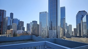 A city skyline with modern high-rise buildings, a clear sky, and a rooftop terrace with a railing.
