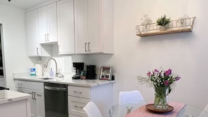 A modern kitchen with white cabinets, a dishwasher, and a coffee maker. There is a glass table with a pink tablecloth and a vase of flowers.