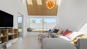 A modern living room with a large flat-screen TV, a wooden bookshelf, a grey sofa with colorful cushions, a small round table, and a large window with blinds. - 3 bedroom lovely home in Gilleleje (Gilleleje)