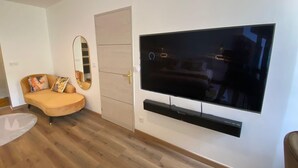 A modern living room with a large flat-screen TV mounted on the wall, a sofa with colorful cushions, a wooden floor, and a door with a mirror. - 19A - The Seventh Heaven, cocoon lover (Tulle)