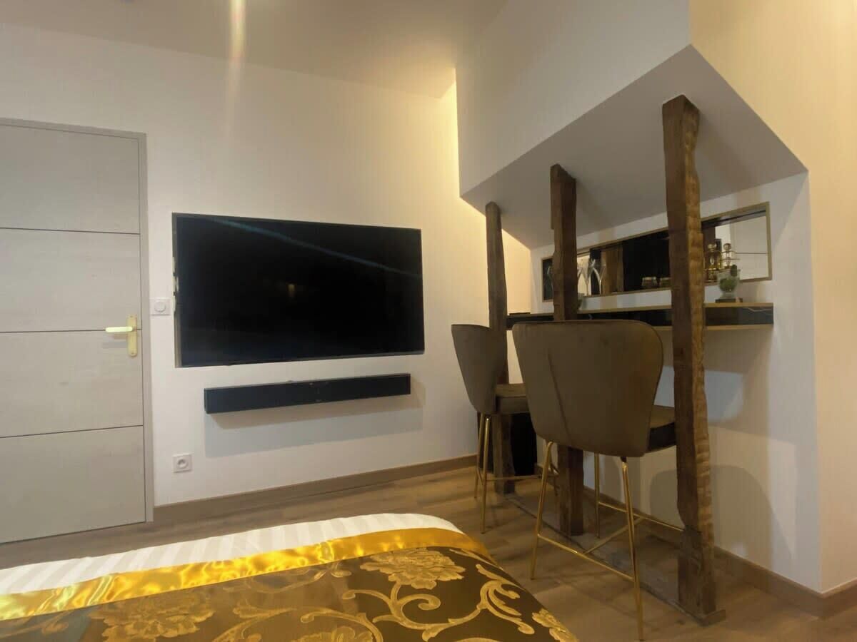 Une chambre à coucher comprenant un lit, un téléviseur fixé au mur, un bureau avec des chaises et une structure en poutres apparentes.