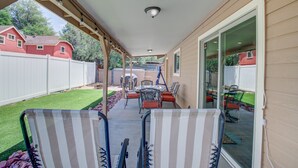 Un patio techado con una mesa y sillas, una cerca blanca y una casa roja al fondo.