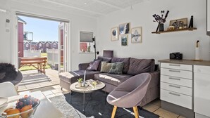 En moderne stue med sofa, sofabord og spiseområde med bænk. Der er billeder i rammer på væggen og en lille hylde med dekorative genstande.