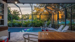 A glass-enclosed pool area with lounge chairs and a table, surrounded by greenery and a clear view of the sky. - Pickleball + Heated Pool | Near Anna Maria (Sarasota)