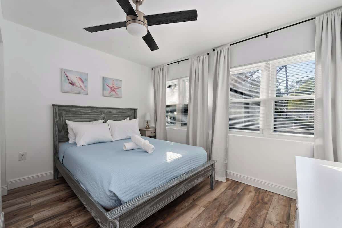 A bedroom with a bed, a ceiling fan, a window with curtains, and artwork on the wall.