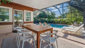A sunlit patio with a pool, wooden table, white chairs, and lounge chairs. - Pickleball + Heated Pool | Near Anna Maria (Sarasota)