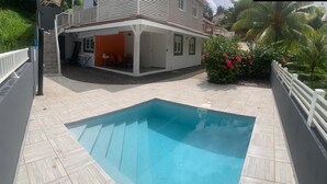 A swimming pool with a wooden deck, a covered patio area, and a view of a house with a balcony. - CINNAMON APPLE (Rivière-Salée)