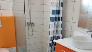 A bathroom with a glass shower enclosure, a white toilet, an orange cabinet, and a white sink with a mirror above it. - CINNAMON APPLE (Rivière-Salée)