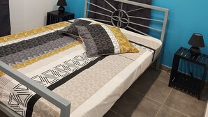 A bed with a metal frame, a headboard with a circular design, and a patterned bedspread. There is a window with a curtain, a wall-mounted air conditioner, and a small table with a lamp. - CINNAMON APPLE (Rivière-Salée)