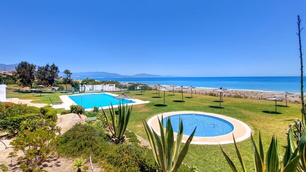A pool area with a rectangular pool and a circular pool, surrounded by grass and plants, with a view of the ocean and mountains in the background.