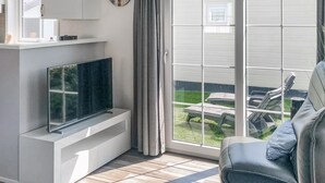Una sala de estar moderna con un televisor de pantalla plana sobre un mueble blanco, un sofá gris y una puerta corrediza de vidrio que da a un patio con mobiliario exterior.