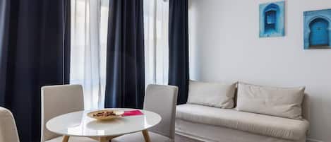 A room with a round table, two chairs, a sofa, and a window with curtains.
