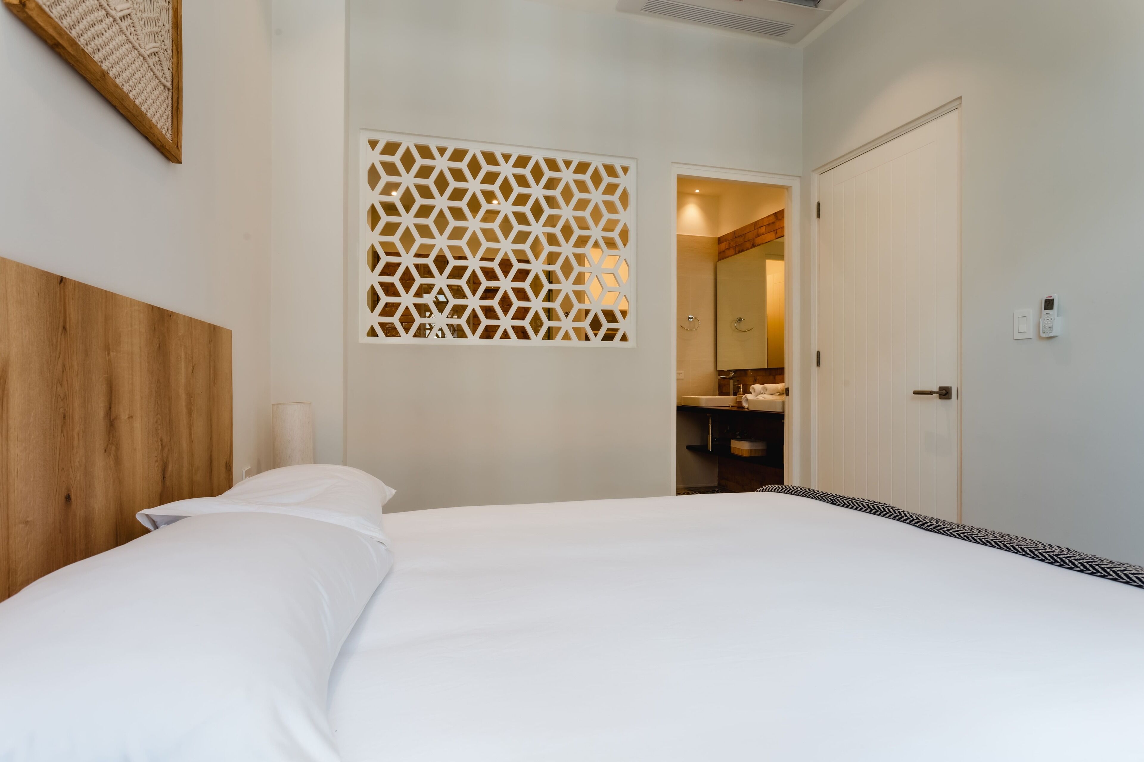 A hotel room with a large bed, a wooden headboard, a decorative wall panel, a bathroom with a mirror, and a white door.