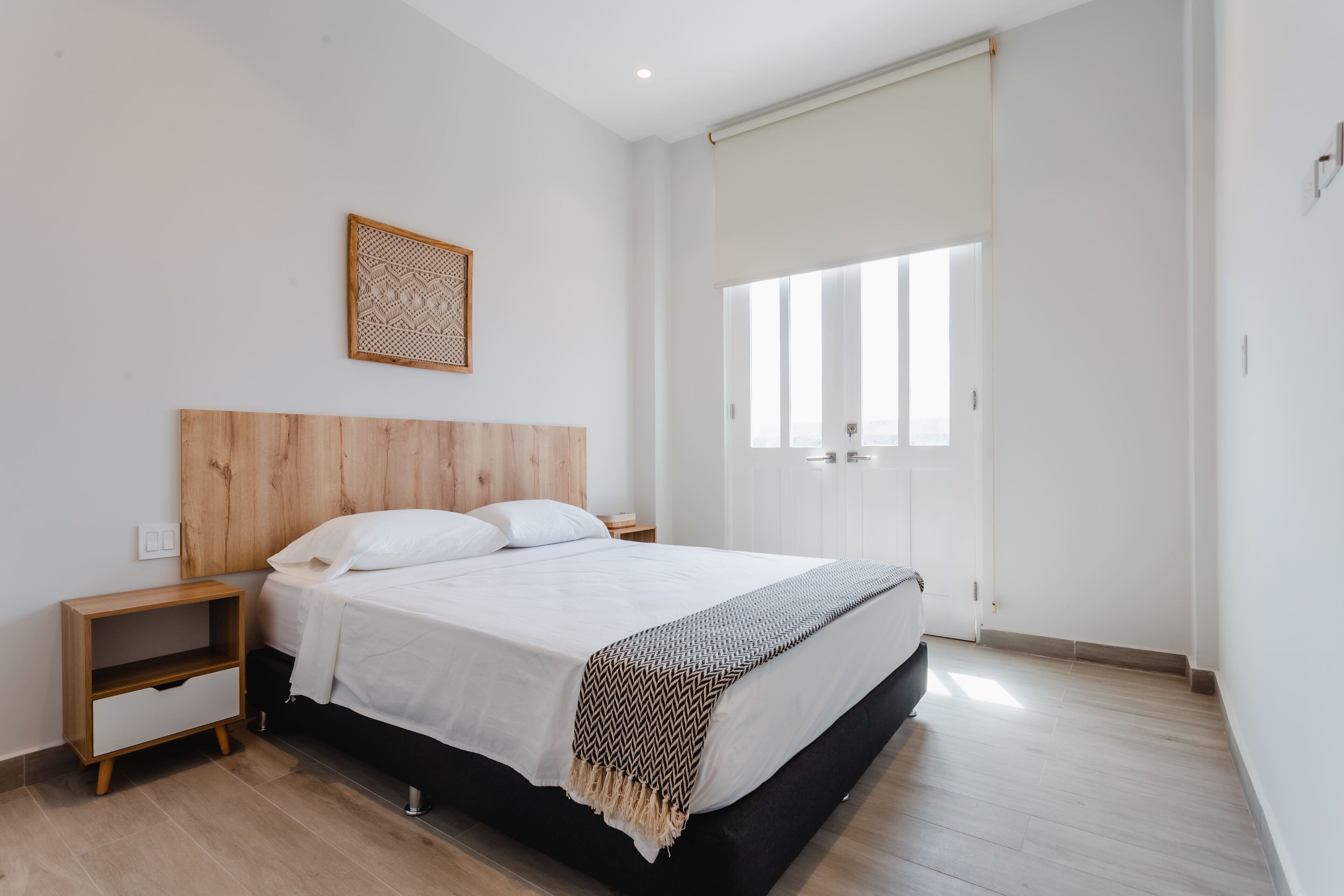 Une chambre moderne avec une tête de lit en bois, un grand lit avec une literie blanche, une table de chevet et une fenêtre avec des stores.