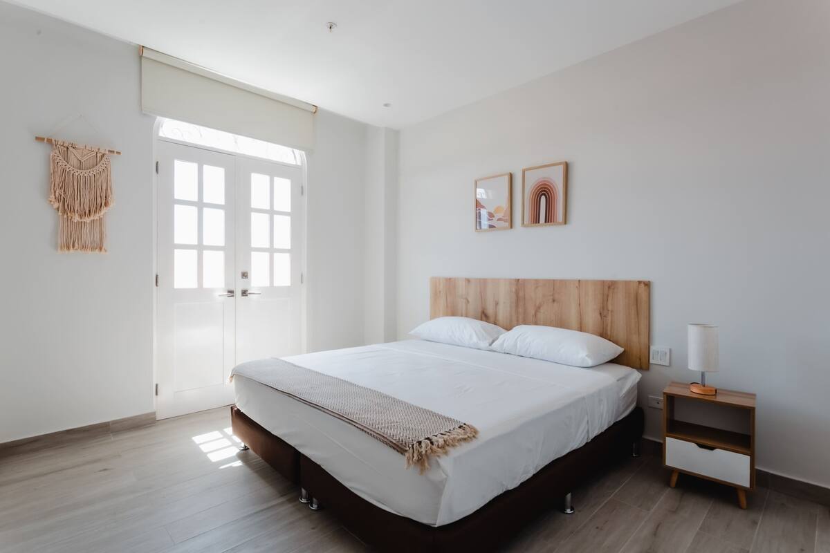 A modern bedroom with a wooden headboard, a large bed with white linens, a bedside table with a lamp, and a macrame wall hanging.