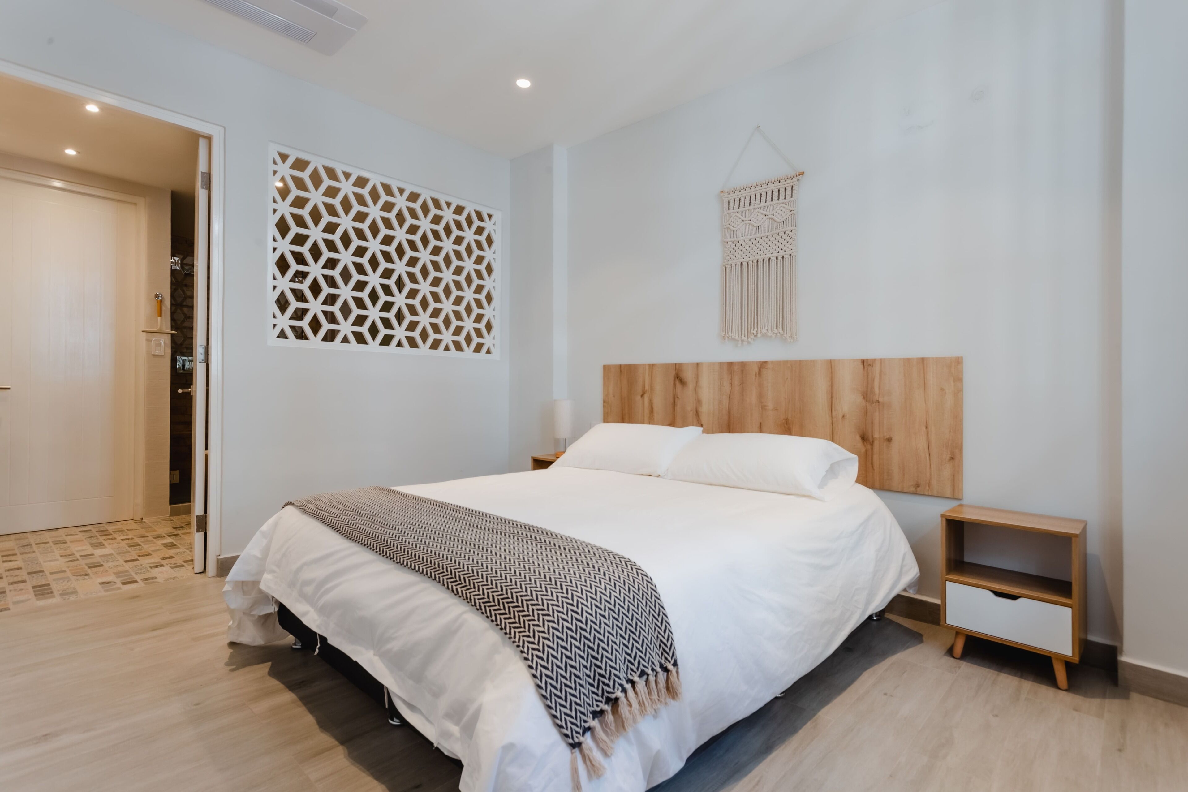 A modern bedroom with a wooden headboard, a bed with white bedding, a patterned throw blanket, a nightstand, and a decorative wall piece.