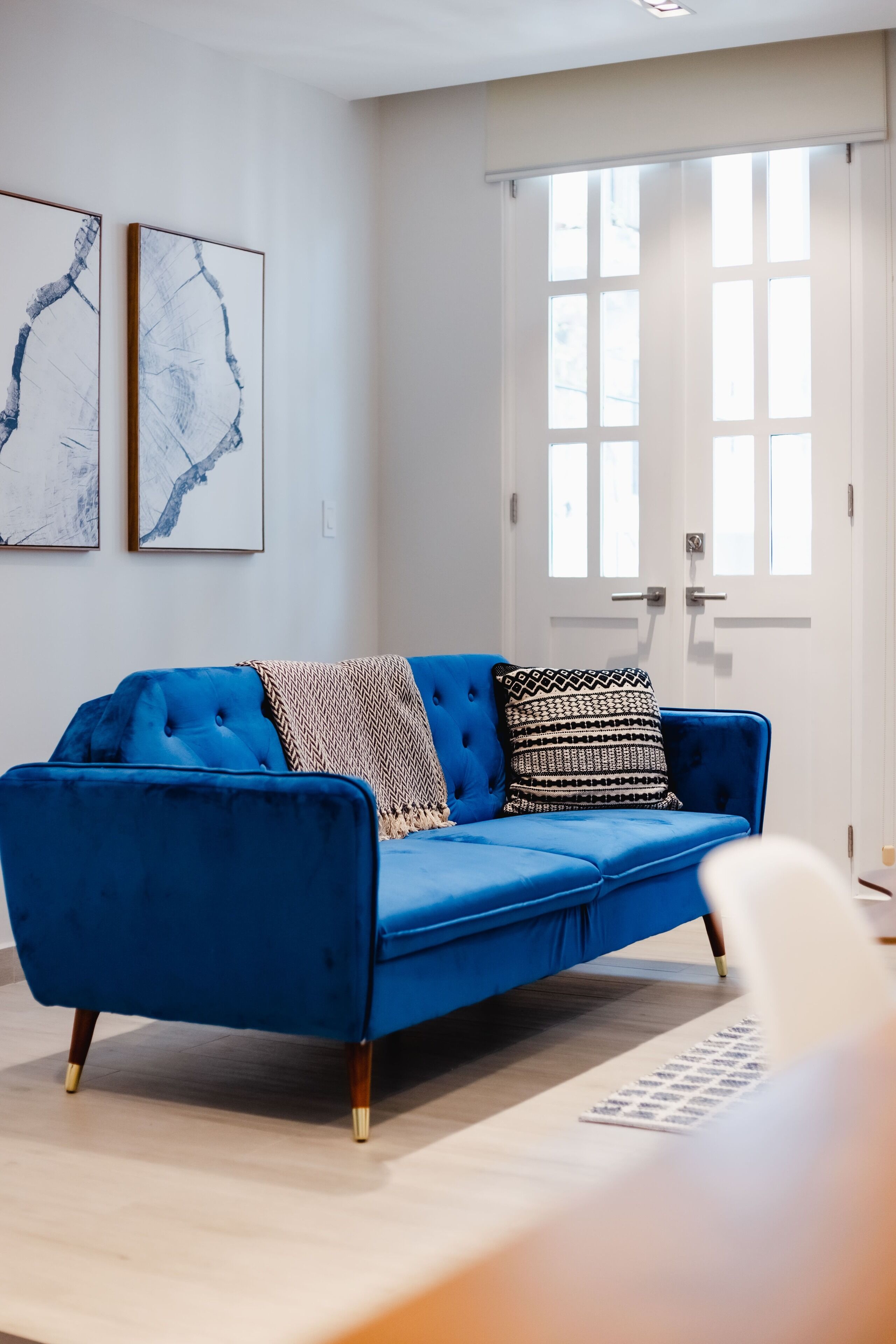 Ein Zimmer mit einem blauen Sofa, zwei abstrakten Bildern, einer weißen Tür und einem Fenster.