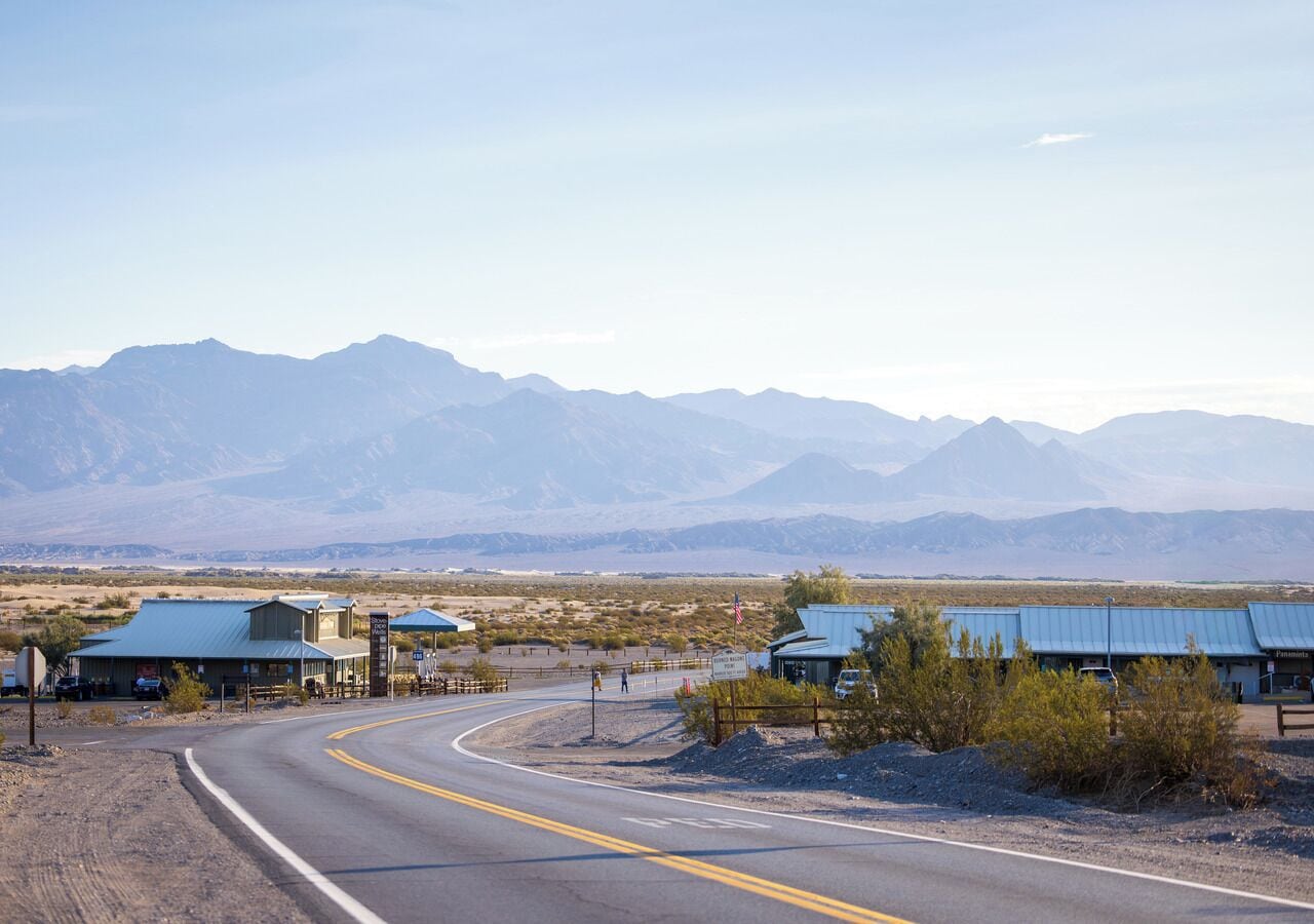 A road curving through a desert landscape with mountains in the background. There are buildings with blue roofs along the roadside.