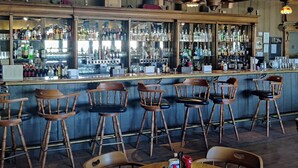 Una zona de bar con sillas de madera y una mesa redonda, estantes llenos de botellas e iluminación colgante.