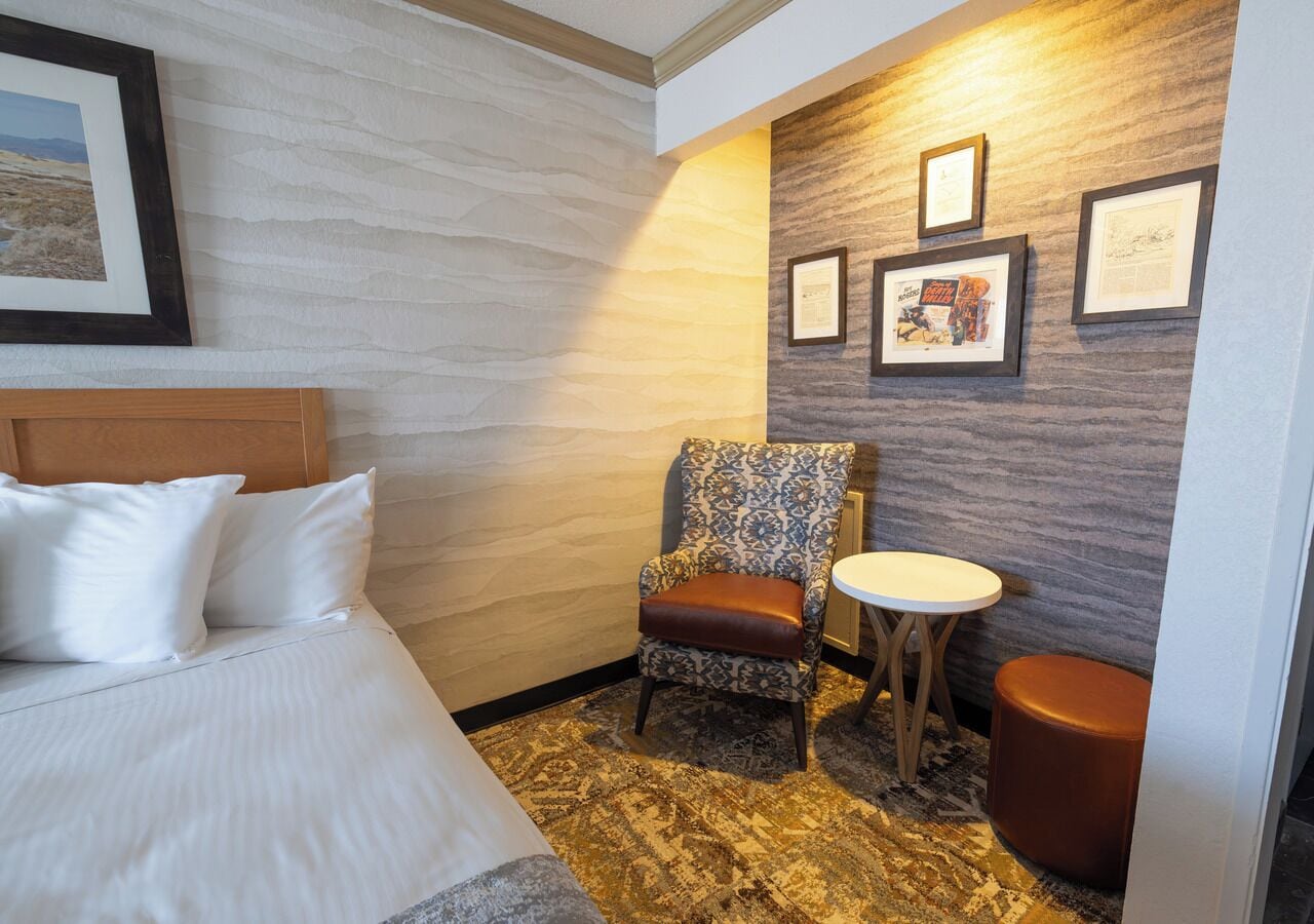 A hotel room with a bed, a patterned armchair, a small round table, and a wall with framed pictures.